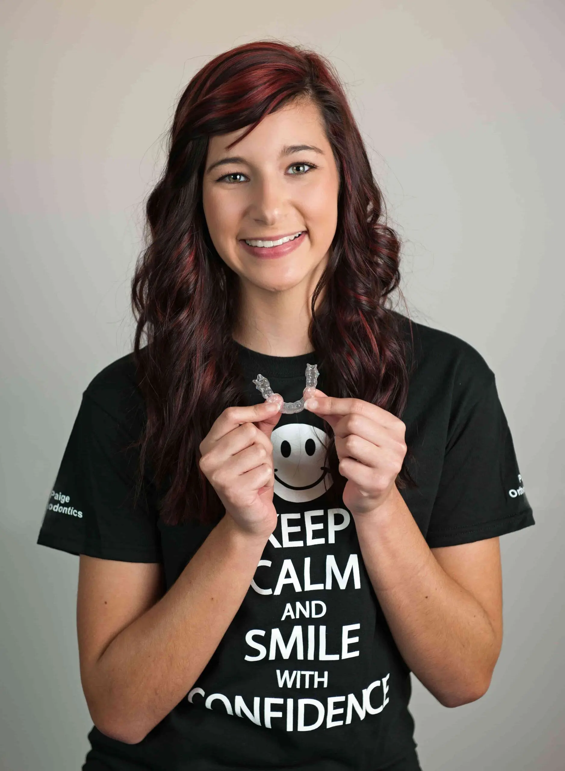 A woman in a black "Keep Calm and Smile" t-shirt holds invisalign, feel to benefits option from an invisalign payment Plan at Paige Orthodontics in Kingwood, TX