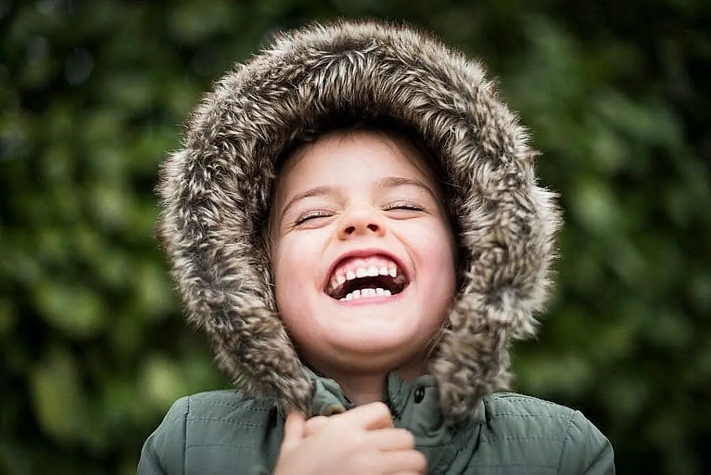 With a blurred green background, a child in a fur-lined hooded coat joyfully showcases their affordable braces with confidence in Kingwood TX.