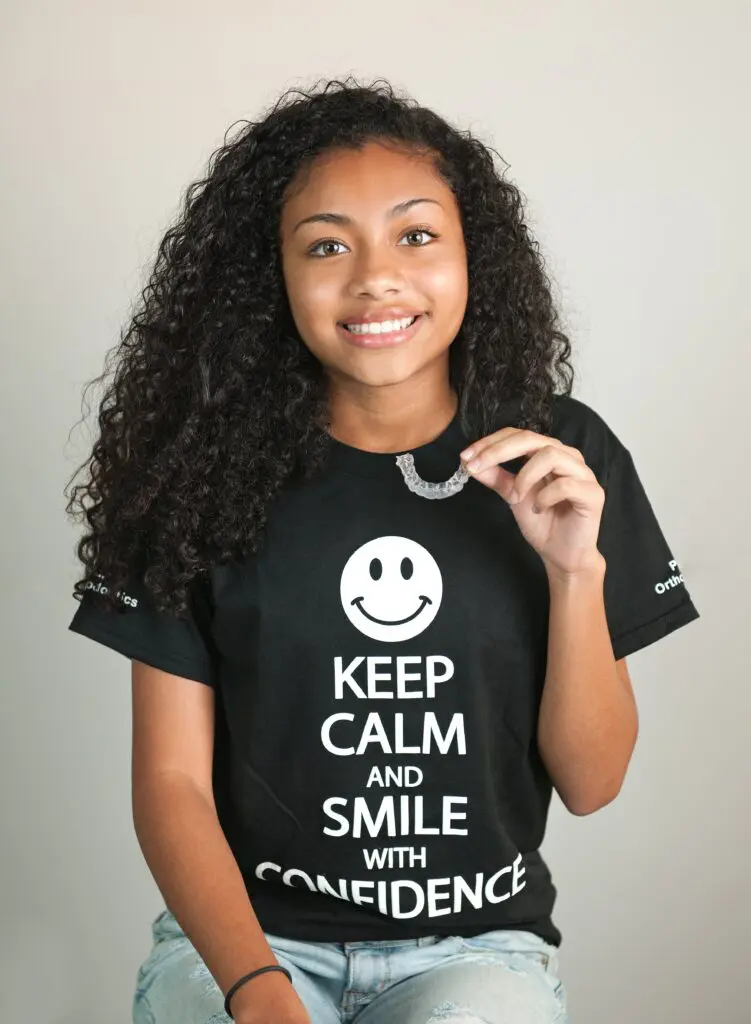 A smiling girl with curly hair, wearing a black shirt that reads "Keep Calm and Smile with Confidence" alongside a smiley face graphic, holds an invisalign clear aligner, as a modern alternatives to braces in Kingwood TX.