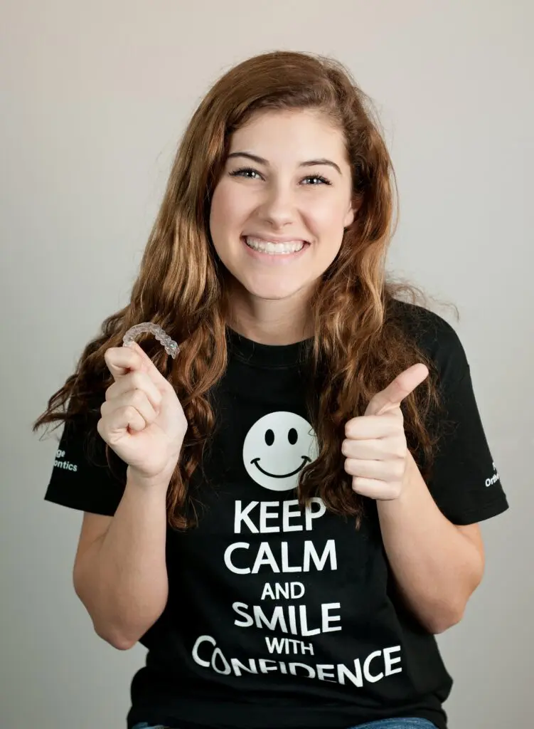 Wearing a "Keep Calm and Smile with Confidence" shirt, a person in Kingwood TX holds an Invisalign clear aligner as one of the alternatives to braces in Kingwood, TX and gives a thumbs-up, smiling at the camera.