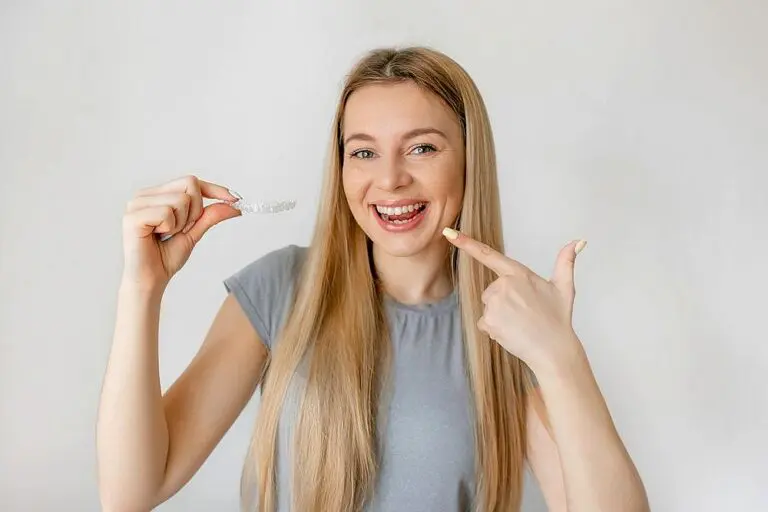 Smiling and pointing at her teeth while holding a clear dental aligner, a young woman in Kingwood TX wonders, "Does insurance cover Invisalign in Kingwood TX?" against a plain light background.