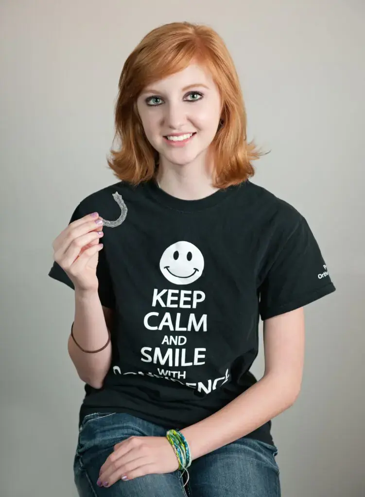 Invisalign vs Braces Cost in Kingwood, TX, a smiling young woman with red hair holds a clear dental aligner while wearing a black t-shirt that reads "KEEP CALM AND SMILE WITH CONFIDENCE. Invisalign vs Braces Cost in Kingwood, TX, a smiling young woman with red hair holds a clear dental aligner while wearing a black t-shirt that reads "KEEP CALM AND SMILE WITH CONFIDENCE.