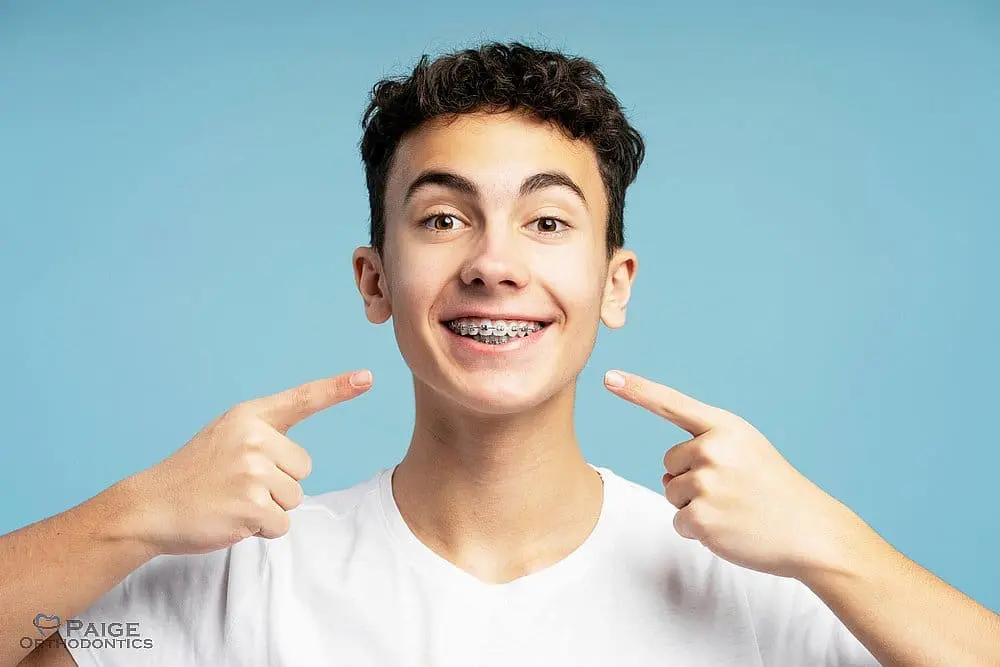 A smiling teenage boy with braces holds dental tools against a light blue background in Kingwood TX - How to Floss with Braces in Kingwood, TX A smiling teenage boy with braces holds dental tools against a light blue background in Kingwood TX - How to Floss with Braces in Kingwood, TX
