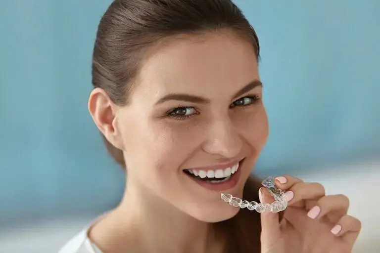 Smiling woman in front of a light blue background holds a clear dental aligner near her mouth in Kingwood TX - Can Invisalign Fix Overbite in Kingwood, TX.