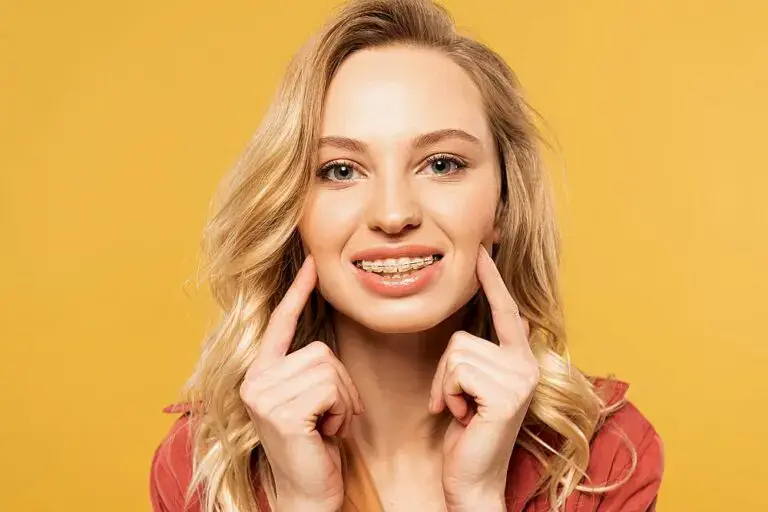 Smiling woman with long blonde hair points to her braces for overbite, standing before a yellow background - Braces for Overbite in Kingwood, TX.