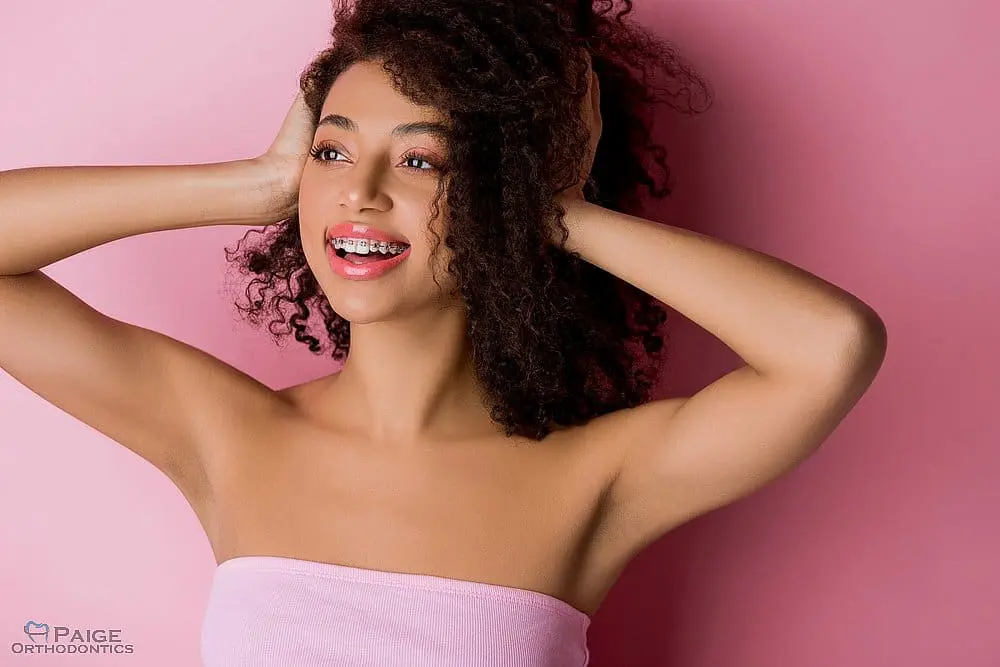 Smiling young woman with curly hair and braces poses in a pink top, hands in hair, pink background—braces glue removal Kingwood TX - How to Get Braces Glue Off Teeth in Kingwood, TX