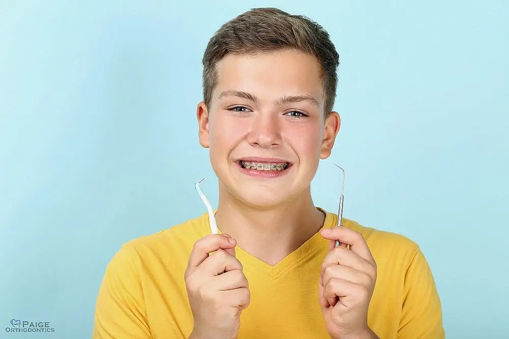 A smiling teenage boy with braces holds dental tools against a light blue background in Kingwood TX - How to Floss with Braces in Kingwood, TX