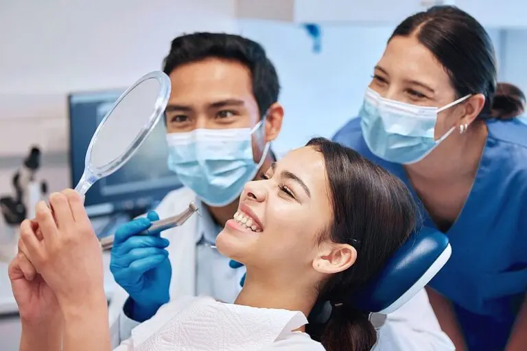 In a Kingwood TX clinic, two dental professionals assist a smiling patient checking her teeth in a mirror after braces glue removal - How to Get Braces Glue Off Teeth in Kingwood, TX