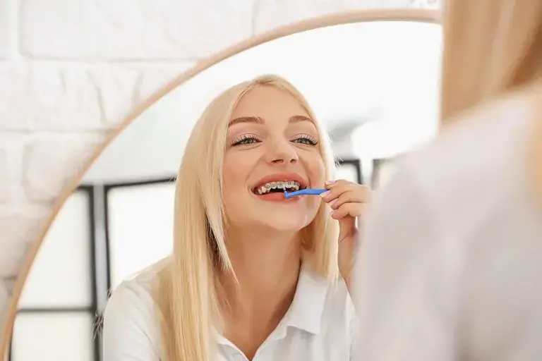 Brushing her teeth in a bathroom mirror, a blonde woman with braces smiles, representing dental care in Kingwood TX - How to Floss with Braces in Kingwood, TX Brushing her teeth in a bathroom mirror, a blonde woman with braces smiles, representing dental care in Kingwood TX - How to Floss with Braces in Kingwood, TX