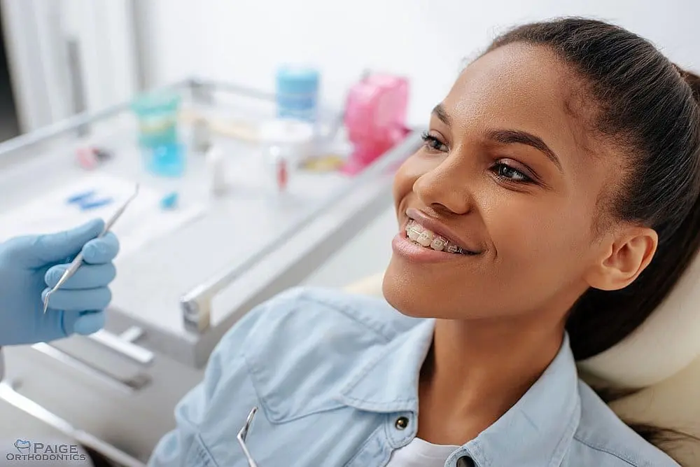 A young woman with braces smiles in a dental chair as a gloved dentist in Kingwood TX holds dental tools nearby - Clear Braces vs Metal Braces in Kingwood, TX 
