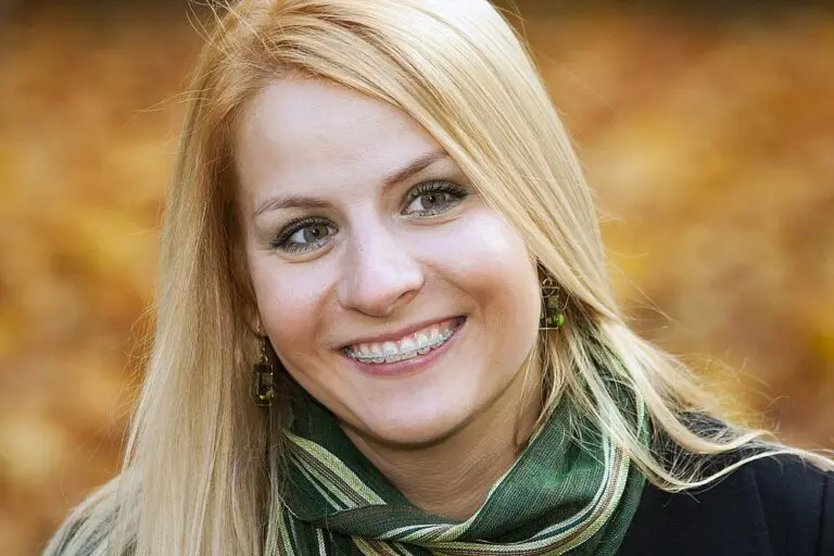 Smiling with braces, a young woman in Kingwood TX wears a green scarf and earrings against a blurred autumn background - Clear Braces vs Metal Braces in Kingwood, TX