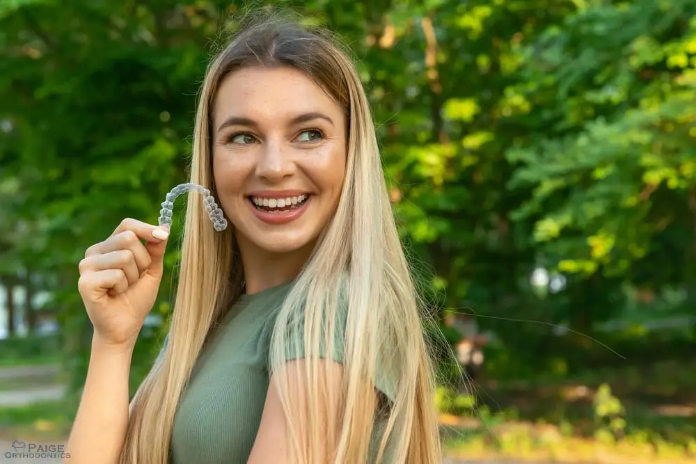 Smiling woman with long blonde hair holds a clear dental aligner outdoors in Kingwood TX, green trees in the background - How Does Invisalign Work in Kingwood, TX