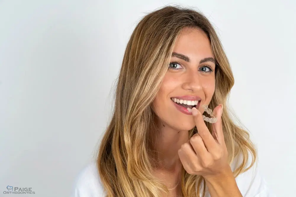 Smiling woman with long blonde hair in Kingwood TX holds a clear orthodontic aligner near her mouth against a white background - How Long Does Invisalign Take in Kingwood, TX 